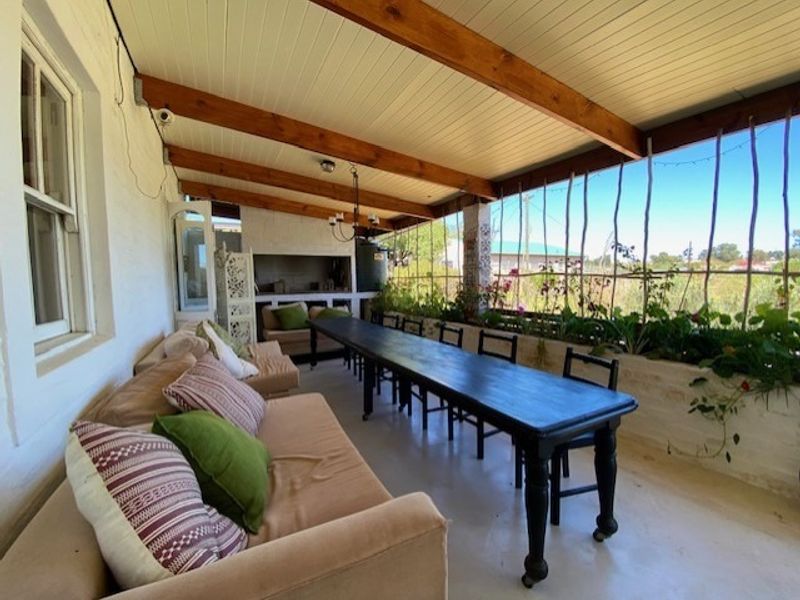 A covered patio featuring exposed timber beams, concrete flooring, and a built-in braai area, overlooking a garden space.