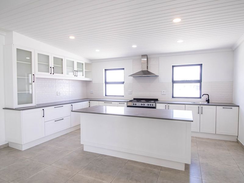 Modern, bright kitchen featuring white cabinetry, a central island, stone countertops, gas stove, and stainless steel extractor fan with tiled flooring.