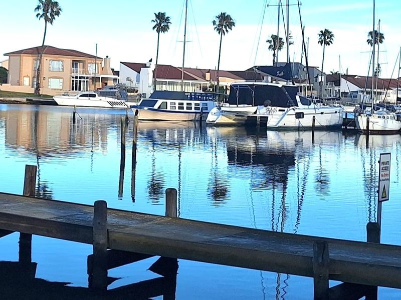 A scenic view of a calm marina with a wooden jetty in the foreground and residential properties in the background.
