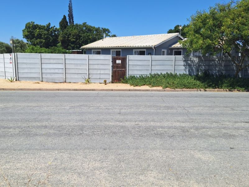 Street view of the property showing the asphalt road, concrete curb, and the precast concrete boundary wall with a pedestrian gate.