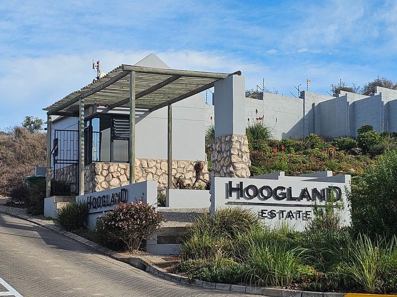 The entrance to Hoogland Estate featuring a stone-clad security gatehouse, perimeter wall with electric fencing, and a paved access road.