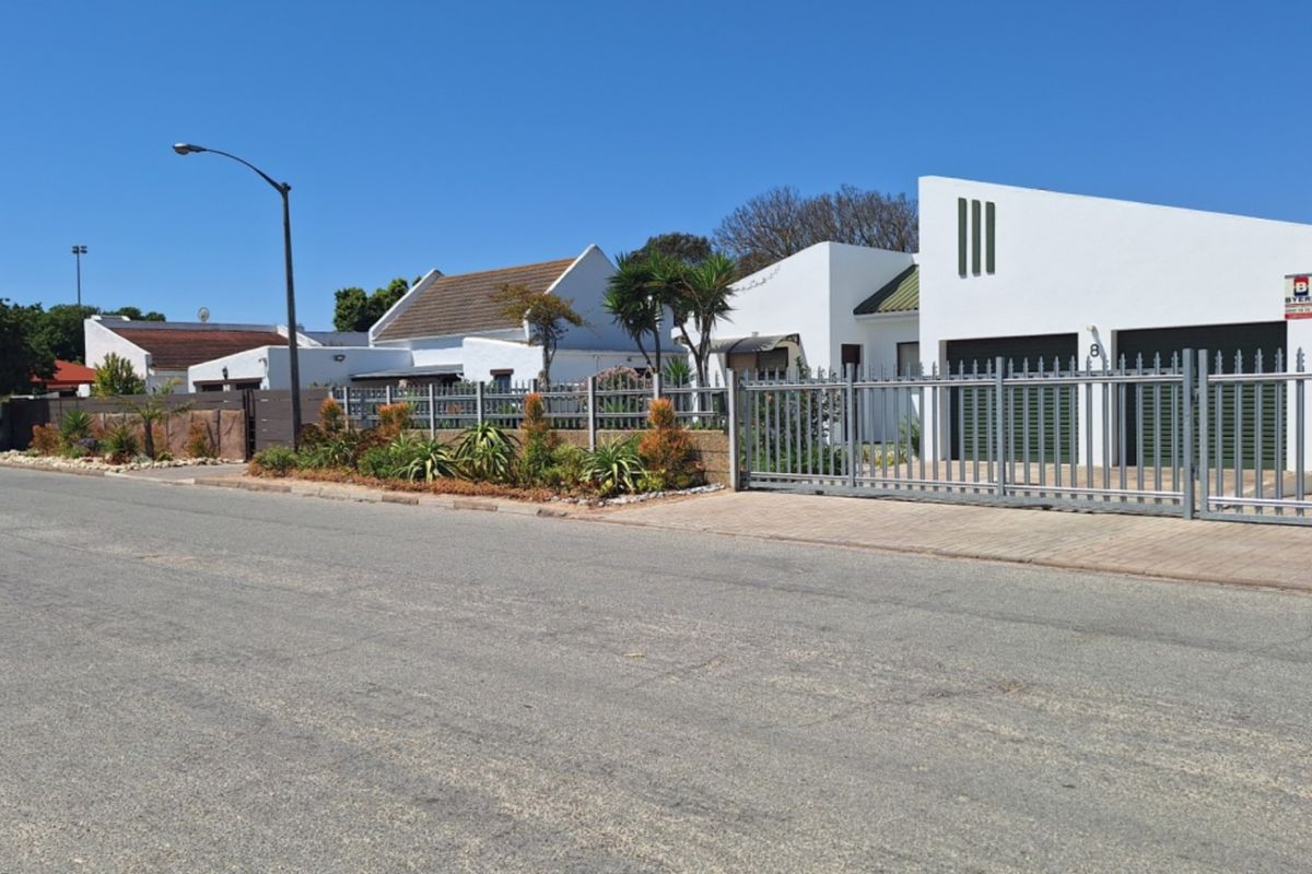 A street-level view of a residential property featuring modern white facades, secure metal fencing, and a paved driveway in a quiet neighborhood.