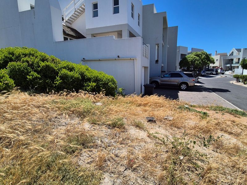 A view of a vacant residential plot adjacent to an existing multi-story residential complex and paved street.