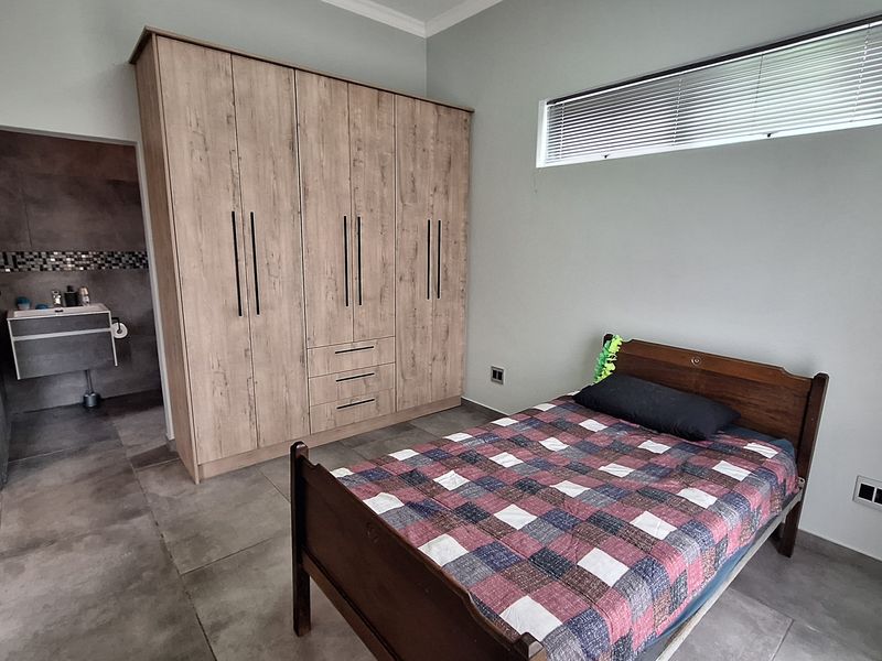 A modern bedroom featuring large format floor tiles, a built-in wooden wardrobe, and a clerestory window, with direct access to an en-suite bathroom.