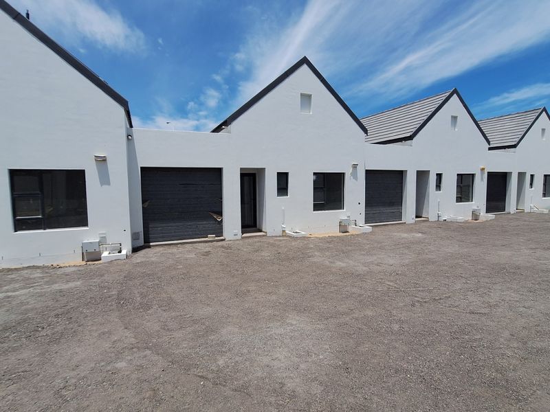 Front elevation of a modern townhouse complex featuring white facades, dark garage doors, and tiled roofs with an unfinished gravel driveway area.