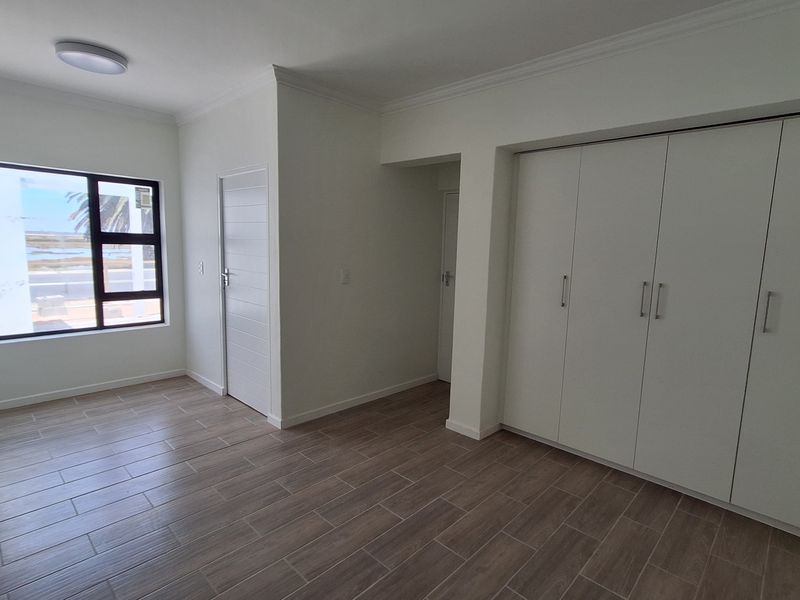 A bright, empty bedroom featuring modern wood-look tiled flooring, built-in cupboards, and a large window providing natural light.