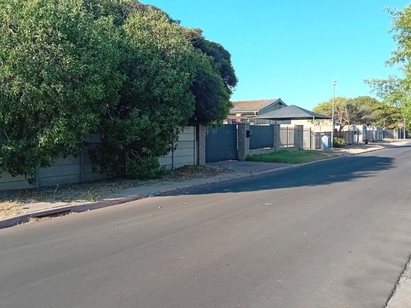 A street-level view of the property frontage, featuring a precast concrete boundary wall, gated driveway access, and a covered carport structure.