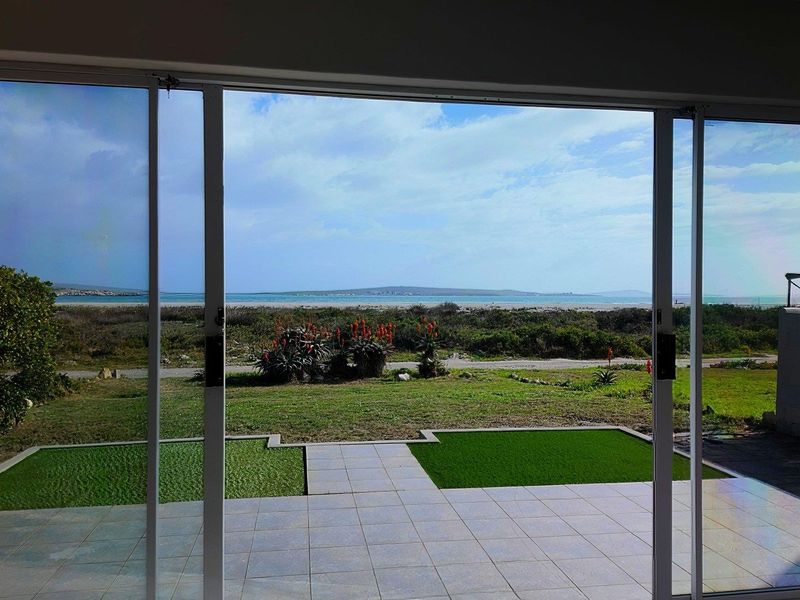 A view from inside looking out through large sliding glass doors onto a tiled patio, artificial lawn, and coastal landscape.