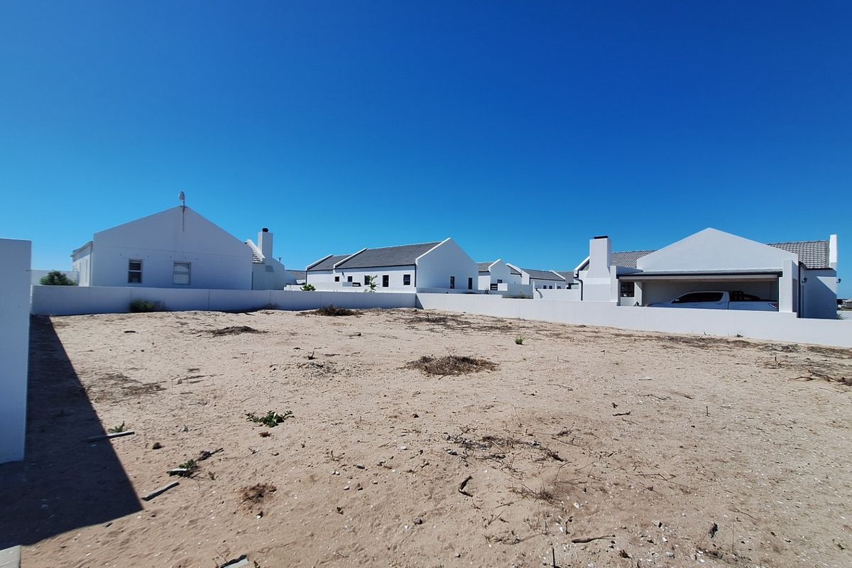 A vacant, sandy plot of land enclosed by a white boundary wall, situated within a residential estate setting.