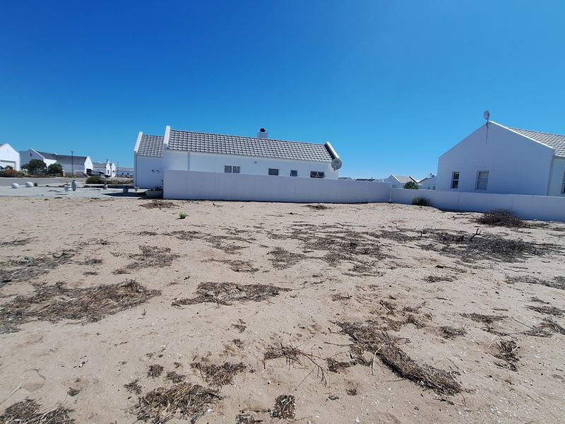 A view of the rear exterior area featuring a sandy, unlandscaped plot enclosed by a white boundary wall with neighboring houses in the background.