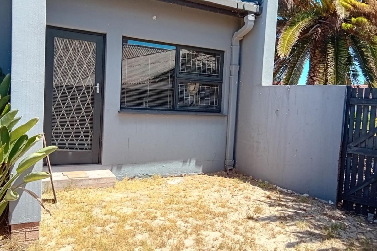 A rear exterior view of the property featuring a security gate, aluminium window, and a small garden area with a timber side gate.