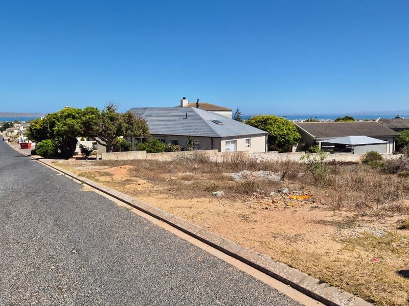 Street view of a vacant residential plot featuring a paved road, concrete curbing, and an existing boundary wall in a coastal neighborhood.