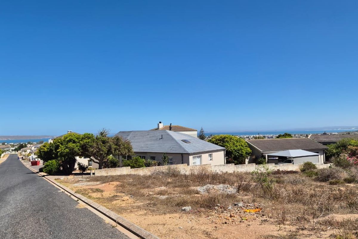 A street-level view of a vacant residential plot featuring a concrete boundary wall, adjacent to an asphalt road with distant coastal views.