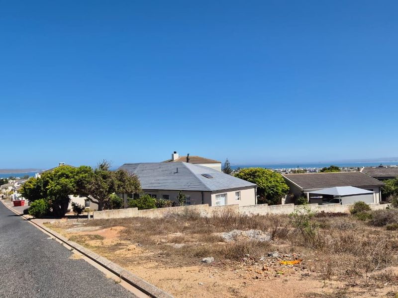 A street-level view of a vacant residential plot featuring a concrete boundary wall, adjacent to an asphalt road with distant coastal views.