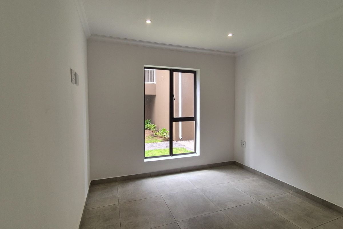 A clean, empty bedroom featuring neutral tiled flooring, recessed downlighting, and a large window providing natural light and a view of the exterior.