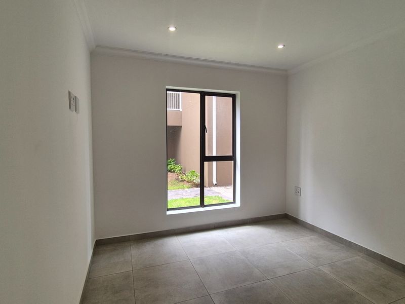 A clean, empty bedroom featuring neutral tiled flooring, recessed downlighting, and a large window providing natural light and a view of the exterior.