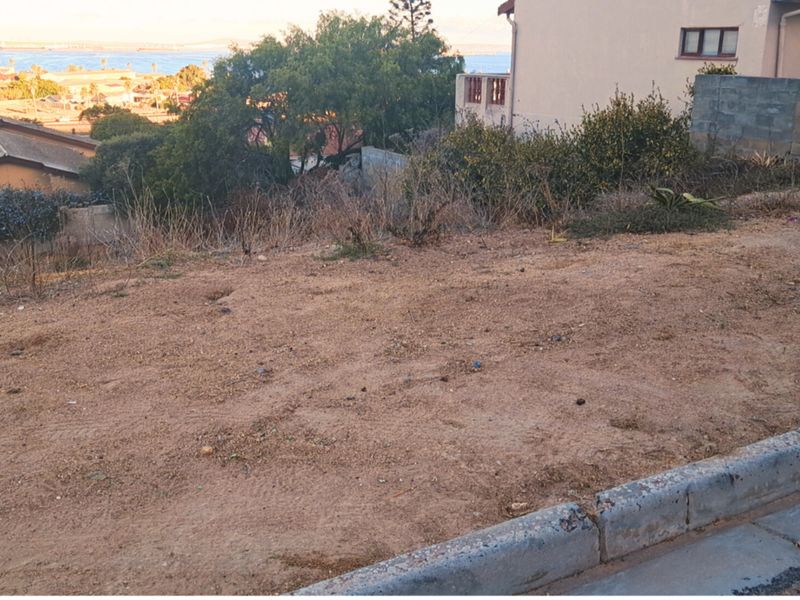 A sloped, unimproved residential vacant lot featuring a concrete curb and distant coastal views.