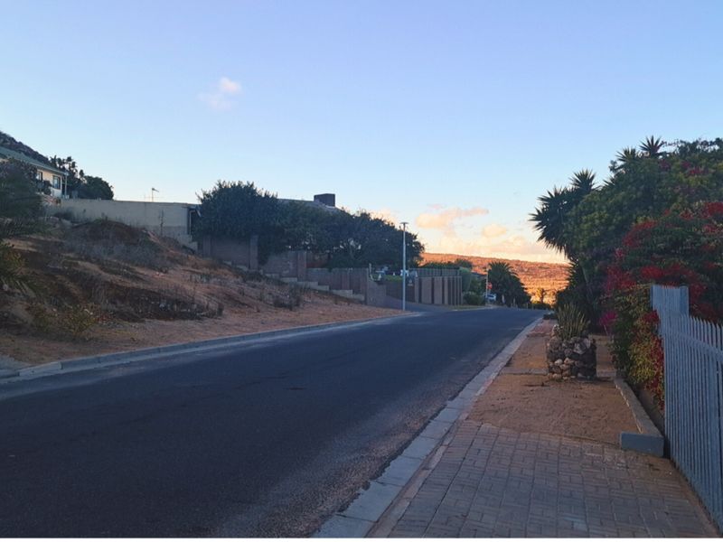 A street-level view of the residential neighborhood, showing the paved road, sidewalk, and surrounding boundary walls and landscaping.