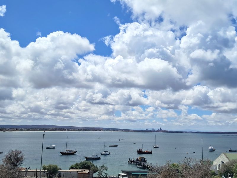 A scenic elevated view overlooking the bay with boats, a small jetty, and distant coastal landscape under a bright blue sky.