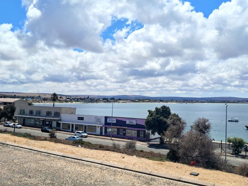 A scenic elevated view overlooking the bay, coastal road, and surrounding commercial buildings under a bright cloudy sky.