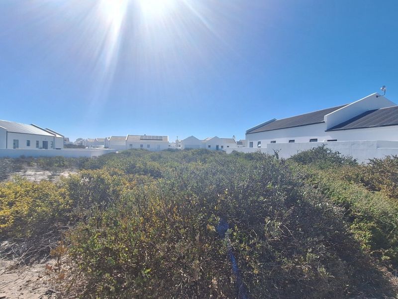 A view of a residential development featuring white buildings with pitched roofs and boundary walls, surrounded by natural coastal vegetation.