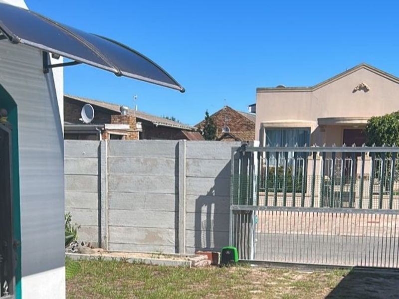 Front exterior view showing a metal sliding gate, precast concrete boundary wall, and a small lawn area with a polycarbonate door awning.