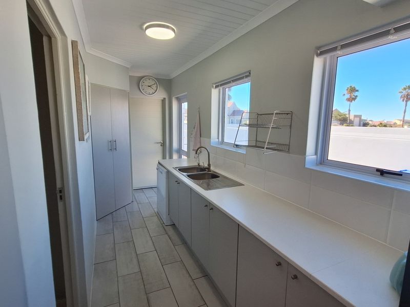A clean and functional scullery featuring built-in cabinetry, a stainless steel double sink, tiled flooring, and ample natural light from large windows.