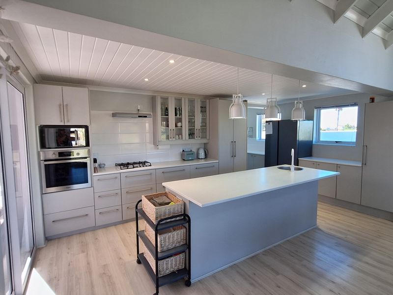 Modern kitchen featuring a central island with sink, built-in oven and microwave, gas hob, and light-toned cabinetry under a vaulted timber ceiling.