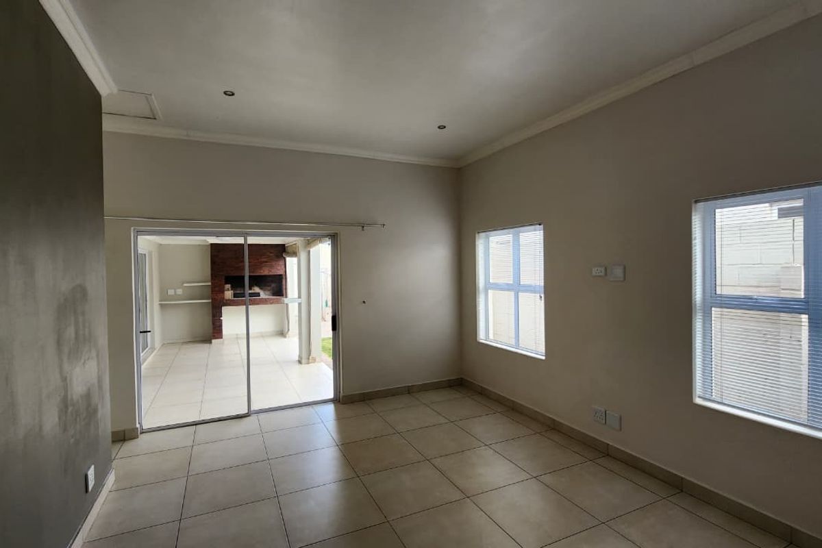 Spacious, light-filled living area featuring neutral tiled flooring, recessed lighting, and a sliding door leading to an outdoor patio with a built-in braai.