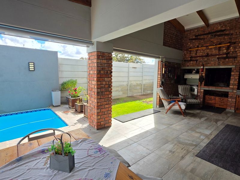 Covered patio area featuring a built-in braai, exposed brickwork, and timber beams, seamlessly connecting to the swimming pool and garden space.