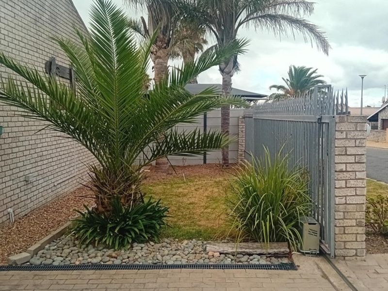 Front exterior view featuring a brick facade, metal security gate, paved driveway, and landscaped garden area with established palm trees.