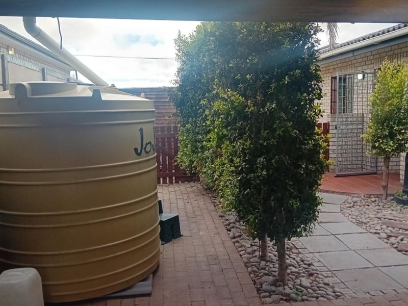 A rear exterior view featuring a large water storage tank, brick paved pathways, and established garden hedging alongside the house structure.