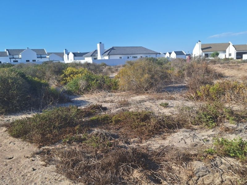 A vacant residential plot featuring sandy terrain and native coastal vegetation, situated in a developed neighborhood with established homes in the background.