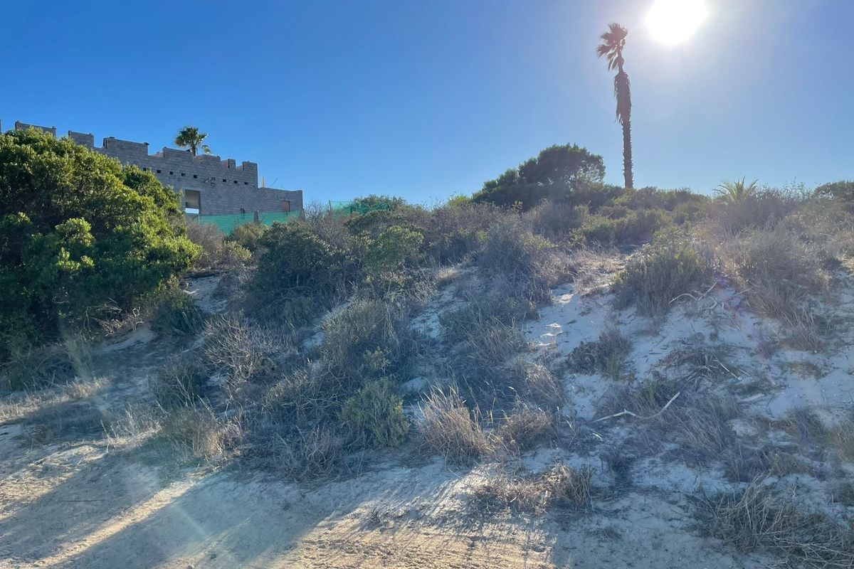 A view of a vacant residential plot featuring sandy terrain, native vegetation, and a partially constructed masonry building in the background.