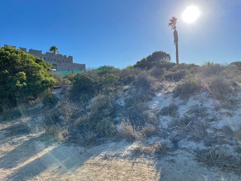 A view of a vacant residential plot featuring sandy terrain, native vegetation, and a partially constructed masonry building in the background.