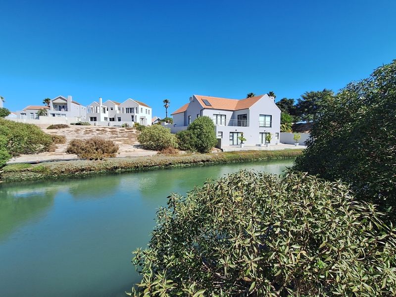 A scenic view of a residential property situated along a tranquil waterway, featuring landscaped surroundings and clear blue skies.