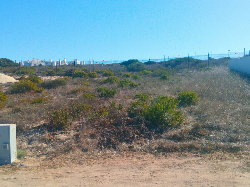 A vacant residential plot featuring natural coastal vegetation, perimeter fencing, and an existing utility connection point.