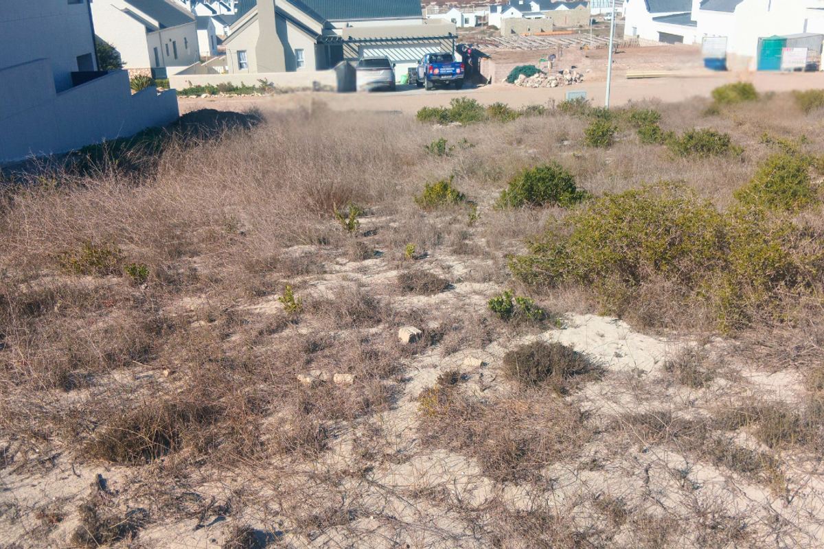 A vacant residential plot featuring sandy soil and native coastal vegetation, situated in a developed neighborhood setting.