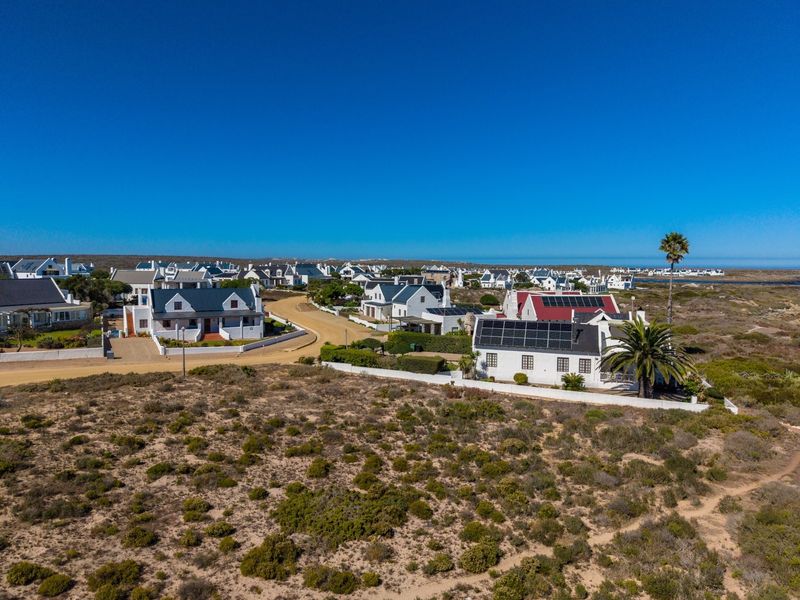 An aerial view of a coastal residential estate featuring white houses with dark roofs, solar installations, and surrounding natural fynbos vegetation.