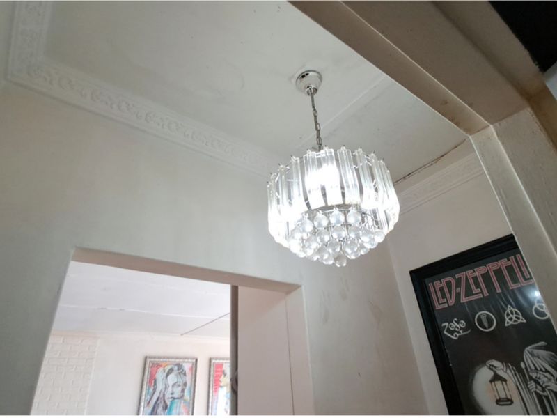 A hallway featuring decorative ceiling cornicing and a crystal chandelier, showing signs of wear including ceiling water marks and wall plaster cracks.