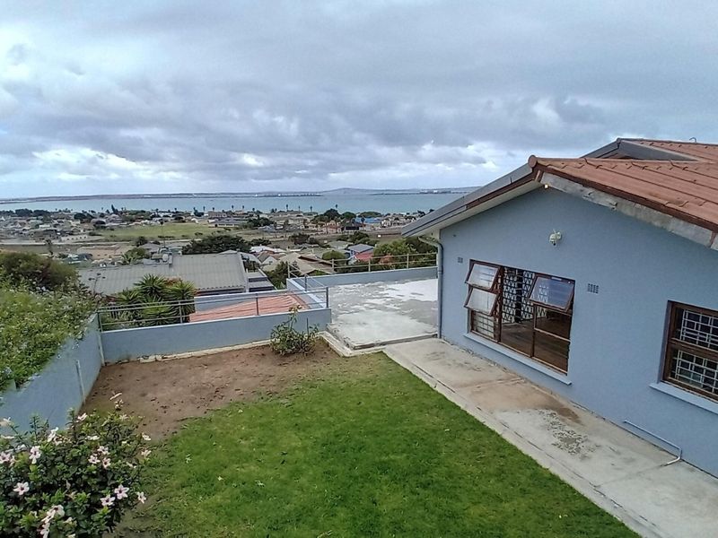 Rear exterior view of the property featuring a lawn, concrete patio, and security-fitted windows, overlooking a coastal town and bay.