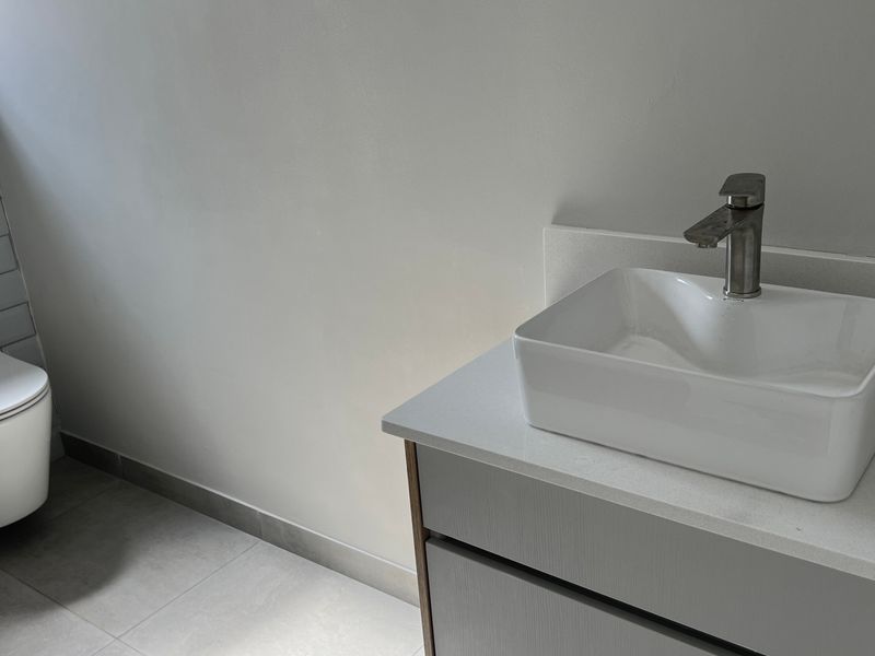 A modern, minimalist bathroom featuring a wall-hung toilet, a sleek vanity cabinet with a countertop basin, and neutral tiled finishes.