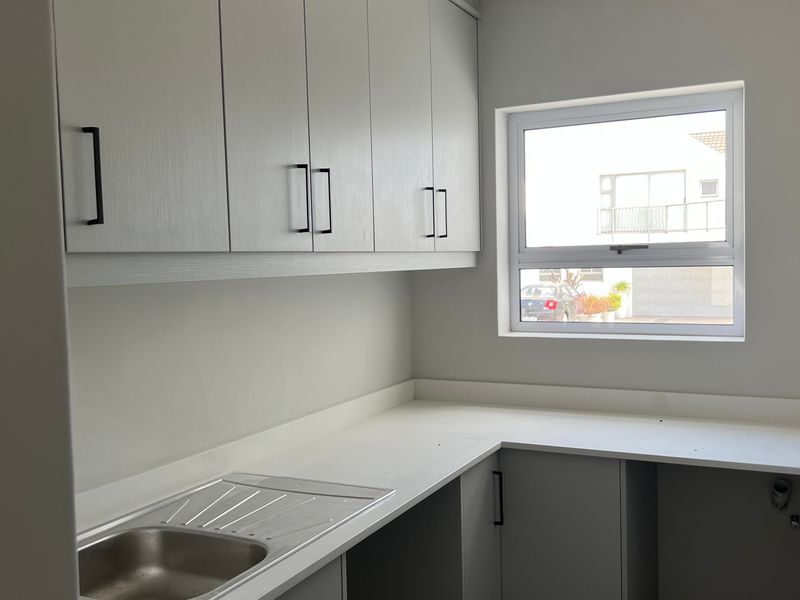 A clean, modern scullery featuring light grey built-in cabinetry, a stainless steel sink, and stone countertops with dedicated plumbing connections.