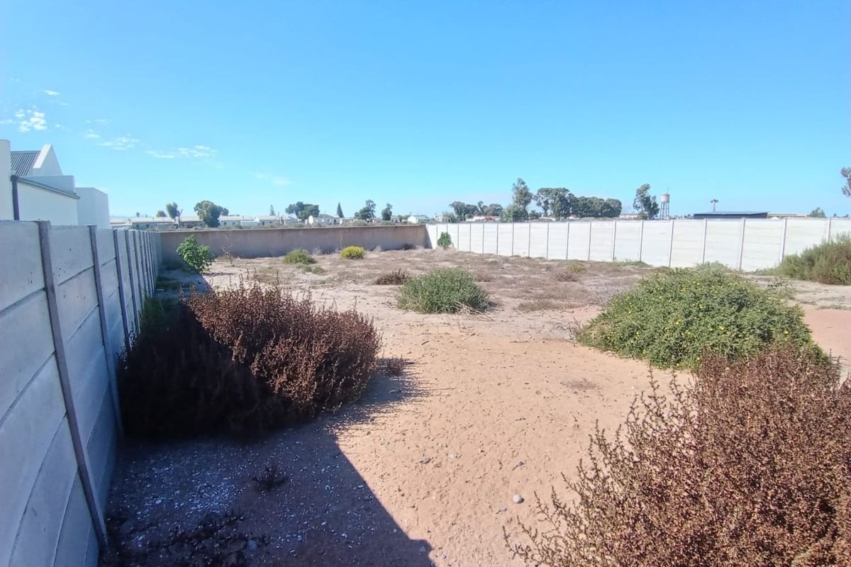 A vacant residential plot enclosed by precast concrete boundary walls, featuring natural sandy ground and low-lying shrubbery.
