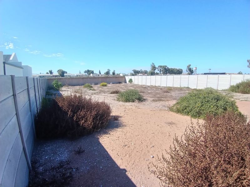 A vacant residential plot enclosed by precast concrete boundary walls, featuring natural sandy ground and low-lying shrubbery.
