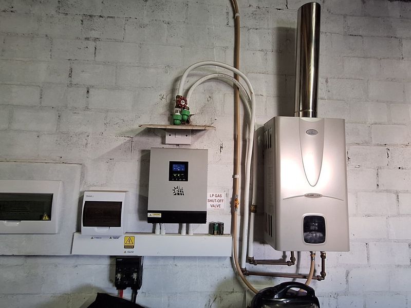 Utility area featuring a wall-mounted gas water heater, solar inverter system, and electrical distribution board against an exposed brick wall.