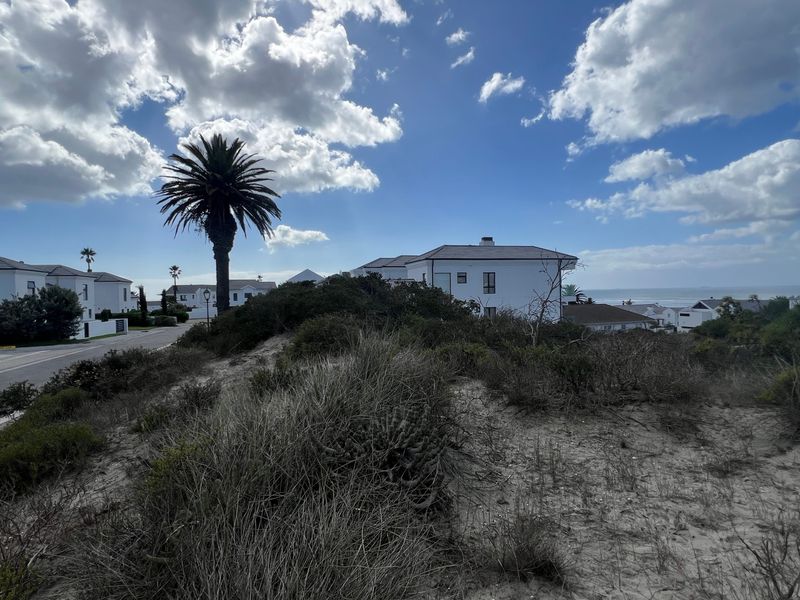 A view of a vacant residential plot featuring natural coastal vegetation, situated in a developed estate with paved road access and distant ocean views.