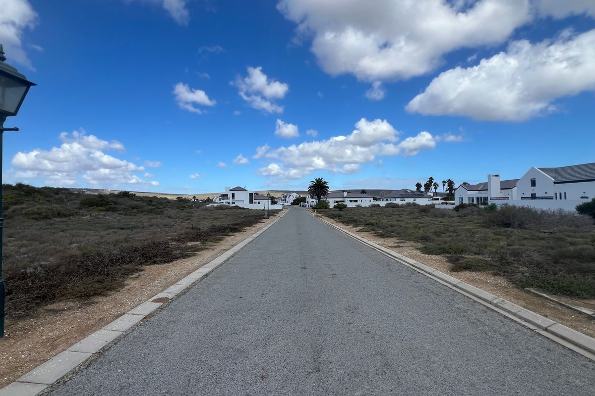 A clear street view showing an asphalt road with concrete curbing and a street lamp, leading towards residential properties in a coastal estate.