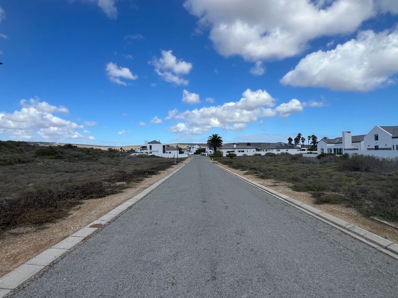 A clear street view showing an asphalt road with concrete curbing and a street lamp, leading towards residential properties in a coastal estate.