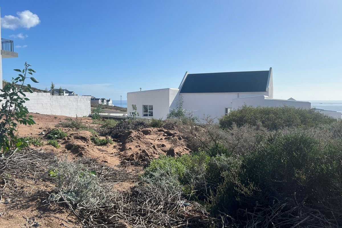A view of a vacant residential plot with natural coastal vegetation, showing adjacent white-walled properties and a glimpse of the ocean.